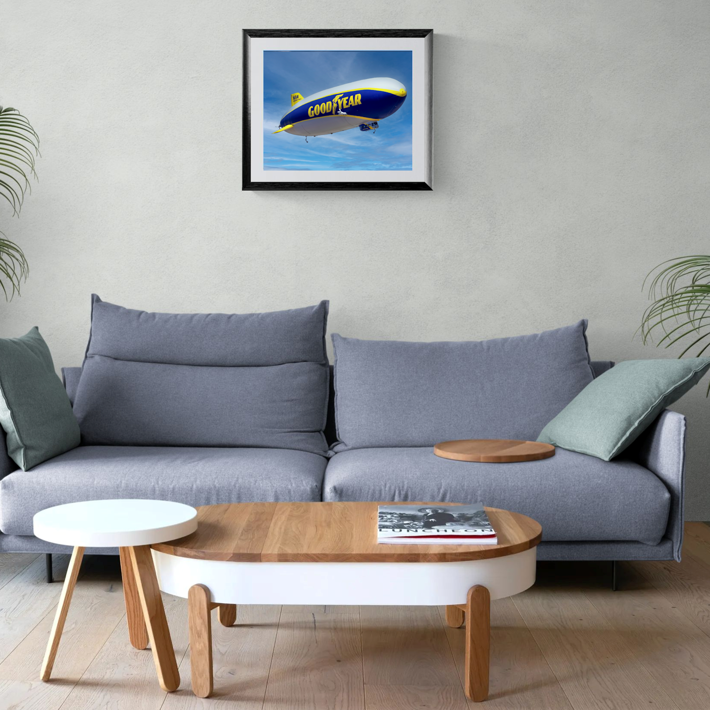 A large-format fine art print of the iconic Goodyear Blimp showcased in a black gallery frame with a clean white mat. The print is centered on a light neutral living room wall directly above a contemporary gray sofa.
