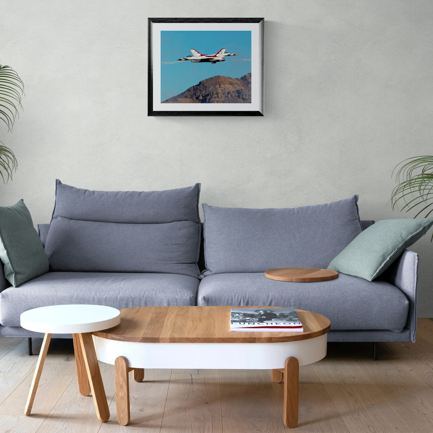 A large-format 16x20 fine art photography print of the USAF Thunderbirds F-16 fighter jets performing a high-speed crossover maneuver against a blue sky and rugged desert mountains. The print is showcased in a black gallery frame with a white mat, centered on a light neutral wall above a contemporary gray sofa with green accent pillows.
