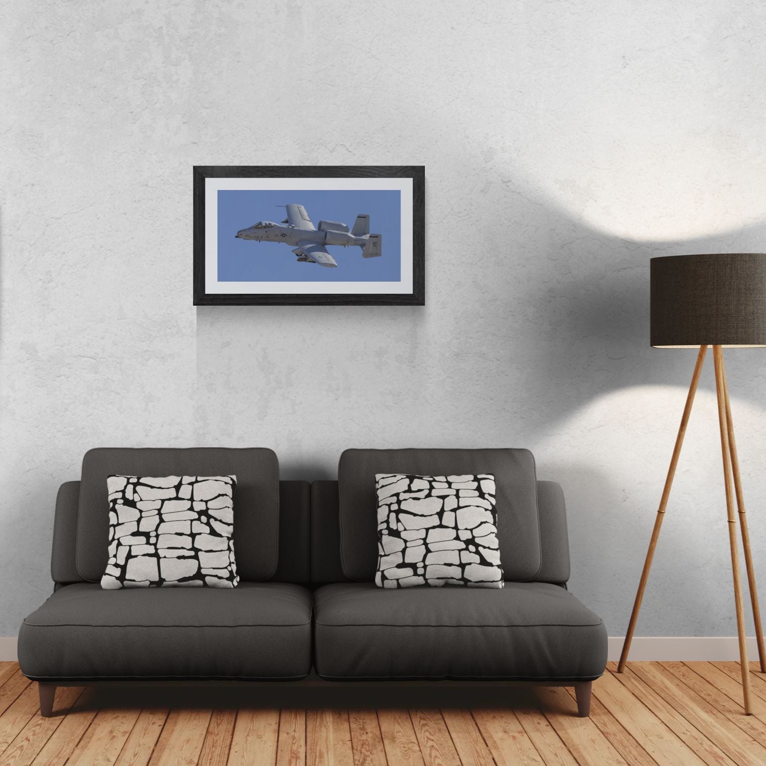 A 30x15 Gallery Edition photograph titled 'A-10 Titanium & Grit,' featuring the rugged, industrial details of an A-10 Thunderbolt II 'Warthog' in a high-fidelity study. The matted aviation print is displayed in a sleek black frame on a light-textured wall above a modern dark gray sofa. A tripod floor lamp stands to the right, providing a precise scale reference for this /50 limited edition residential deployment.