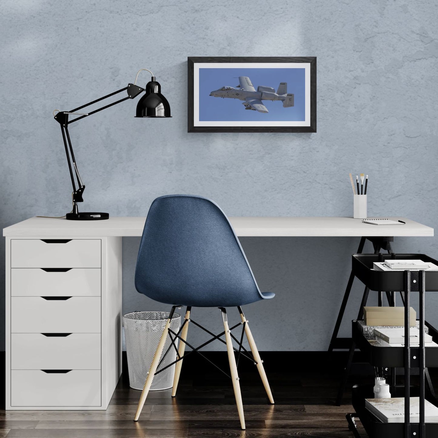 A 20x10 "Standard Edition" of the "A-10 Thunderbolt II: Titanium & Grit" print featured in a modern home office. The textured black gallery frame is mounted on a light-blue wall above a white desk, illustrating the panoramic scale and tactical detail of the Warthog in a personal workspace.