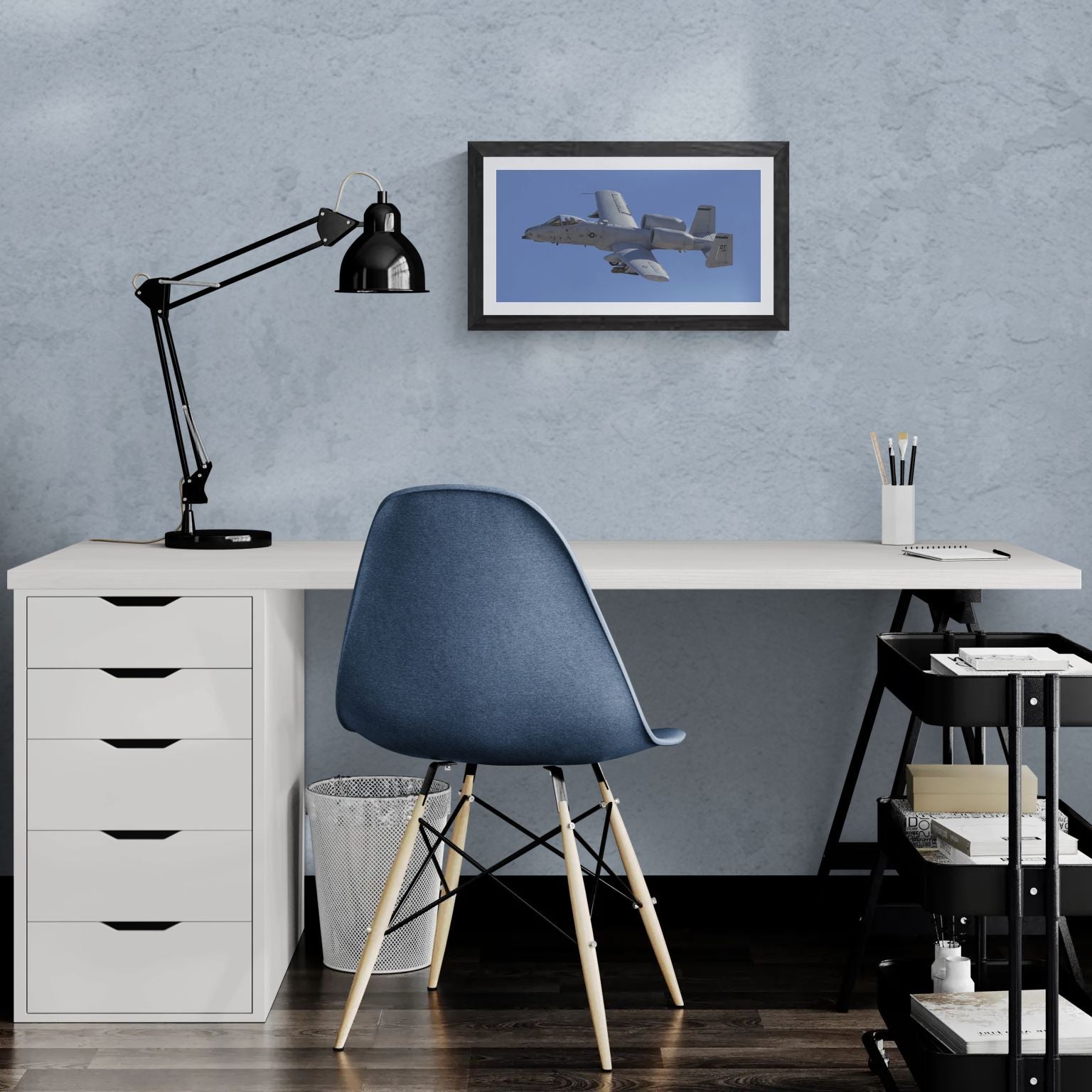A 20x10 "Standard Edition" of the "A-10 Thunderbolt II: Titanium & Grit" print featured in a modern home office. The textured black gallery frame is mounted on a light-blue wall above a white desk, illustrating the panoramic scale and tactical detail of the Warthog in a personal workspace.