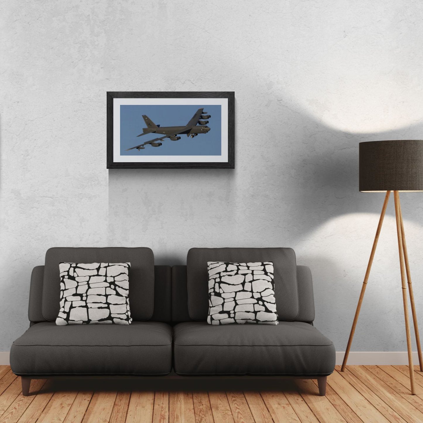 A 30x15 Gallery Edition aviation photograph titled 'B-52 Industrial Titan,' capturing the massive, monolithic profile of a B-52 Stratofortress in a high-detail industrial study. The matted print is professionally displayed in a black gallery frame on a light-textured wall above a modern dark gray sofa. A tripod floor lamp to the right provides a residential scale reference for this /50 limited edition deployment.