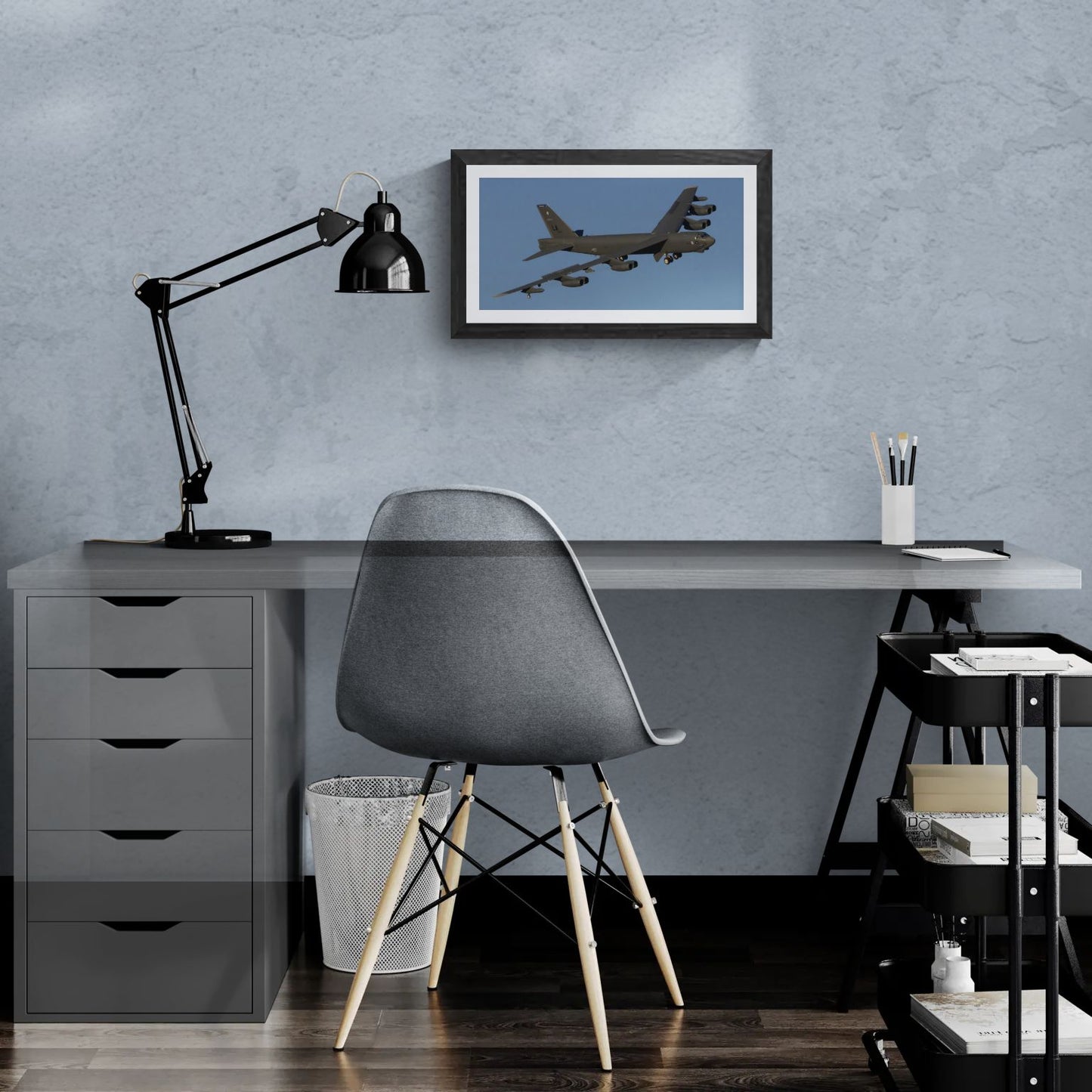 A 20x10 "Standard Edition" of the "B-52 Stratofortress: The Industrial Titan" print featured in a modern home office. The textured black gallery frame is mounted on a light-blue textured wall above a white desk, illustrating the panoramic scale and industrial detail of the B-52 in a personal workspace.