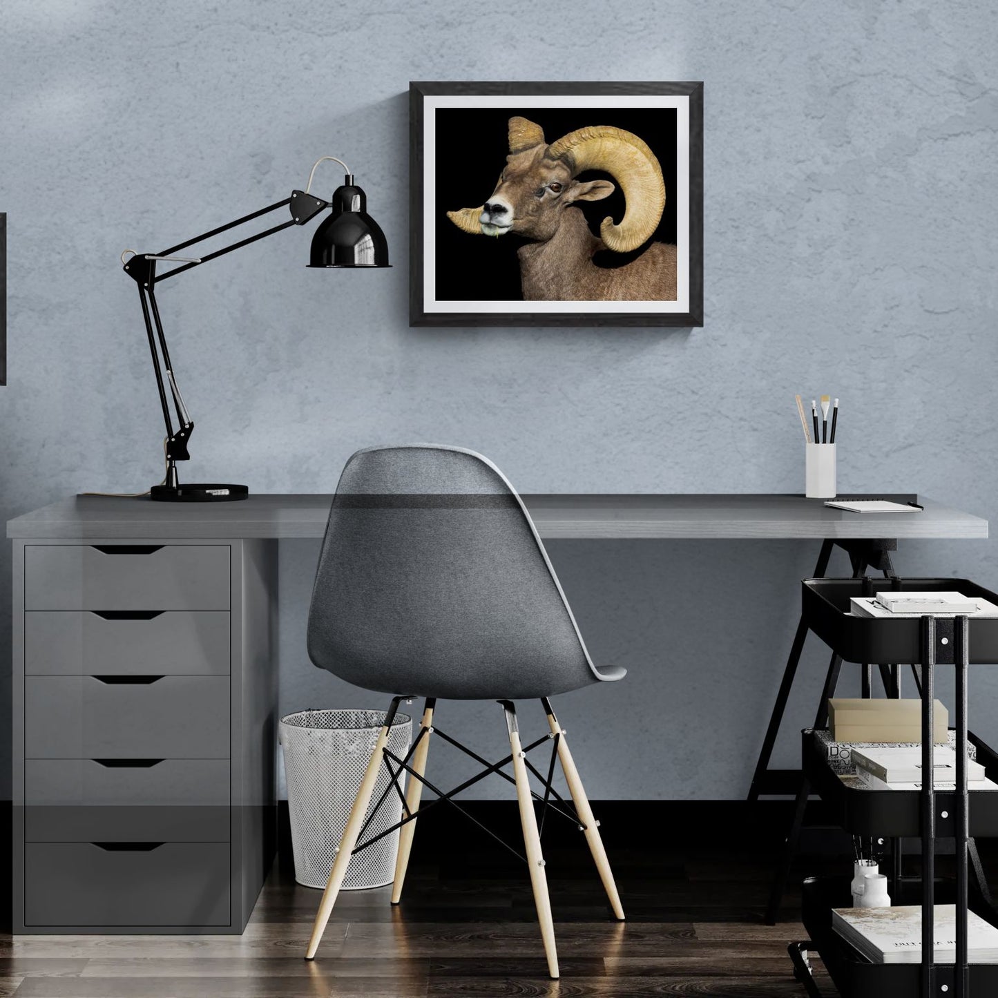 A 20x16 "Standard Edition" of the "Desert Monarch" Bighorn Ram print featured in a modern home office. The textured black gallery frame anchors the wildlife portrait on a light-blue textured wall above a white desk, illustrating the print’s biological detail and scale for personal galleries.