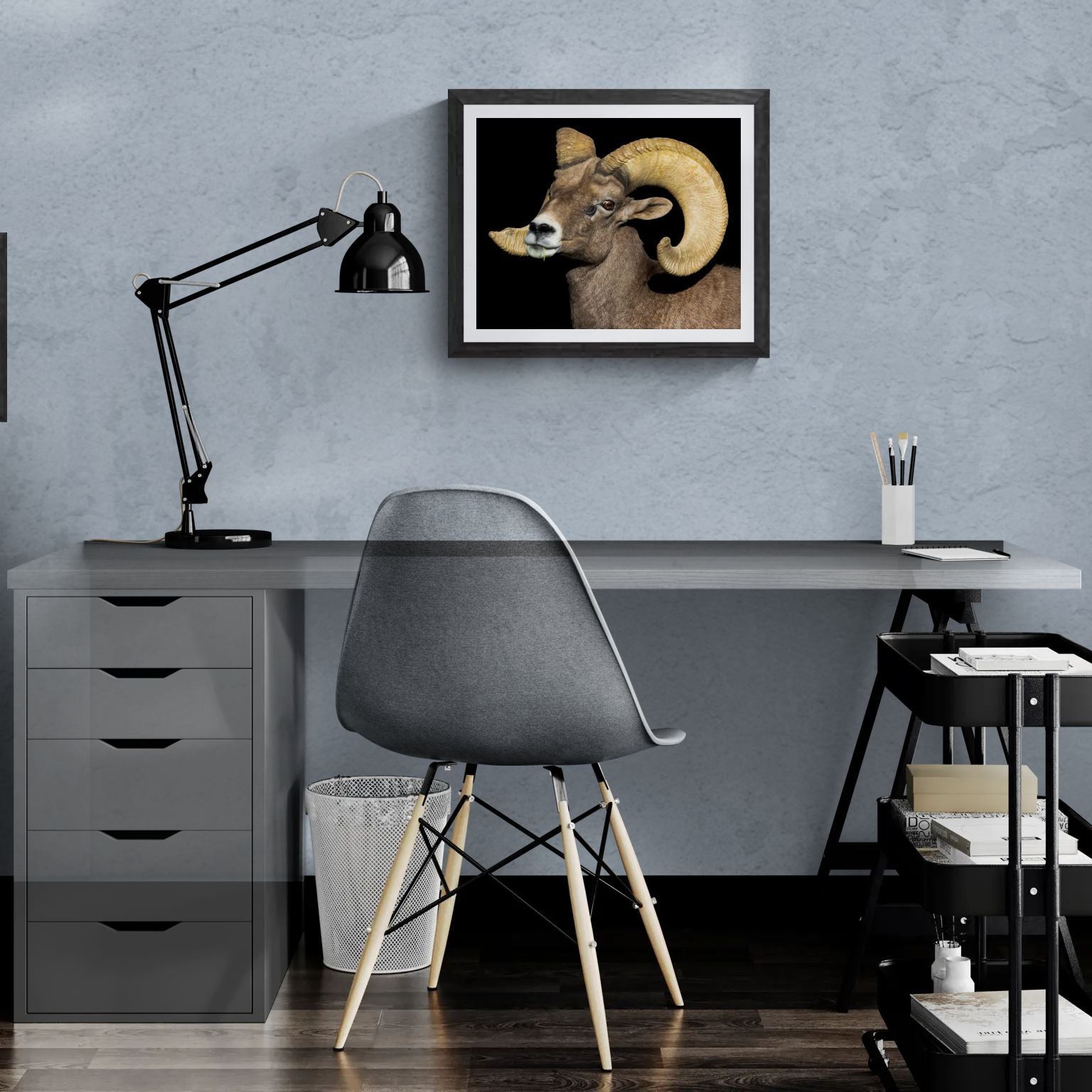 A 20x16 "Standard Edition" of the "Desert Monarch" Bighorn Ram print featured in a modern home office. The textured black gallery frame anchors the wildlife portrait on a light-blue textured wall above a white desk, illustrating the print’s biological detail and scale for personal galleries.