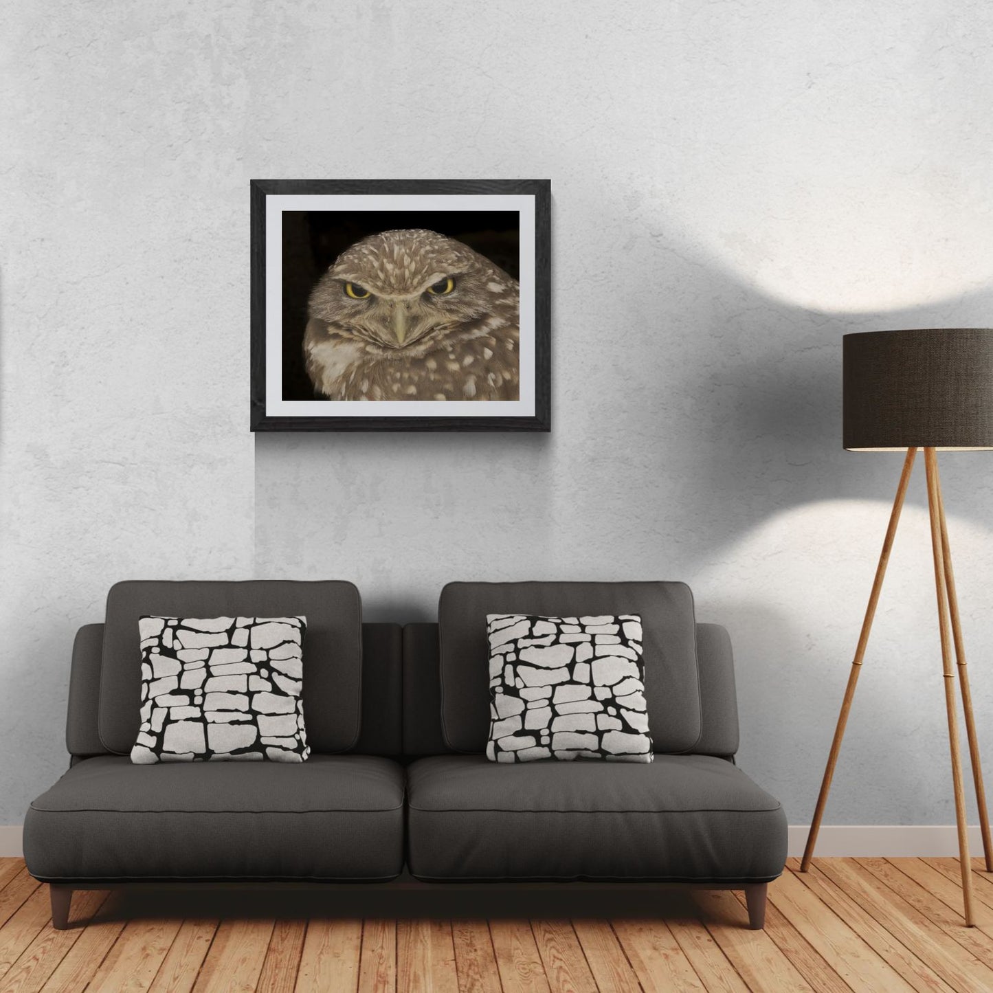 A 30x24 'Gallery Edition' of the 'Piercing Intensity' wildlife print, featuring a close-up study of a Burrowing Owl with vivid yellow eyes. The textured black gallery frame anchors the matted print on a light-textured wall above a modern dark gray sofa. To the right, a tripod floor lamp provides a residential scale reference for this /50 limited edition deployment, highlighting the sharp detail and compelling gaze of the subject for a primary living space.