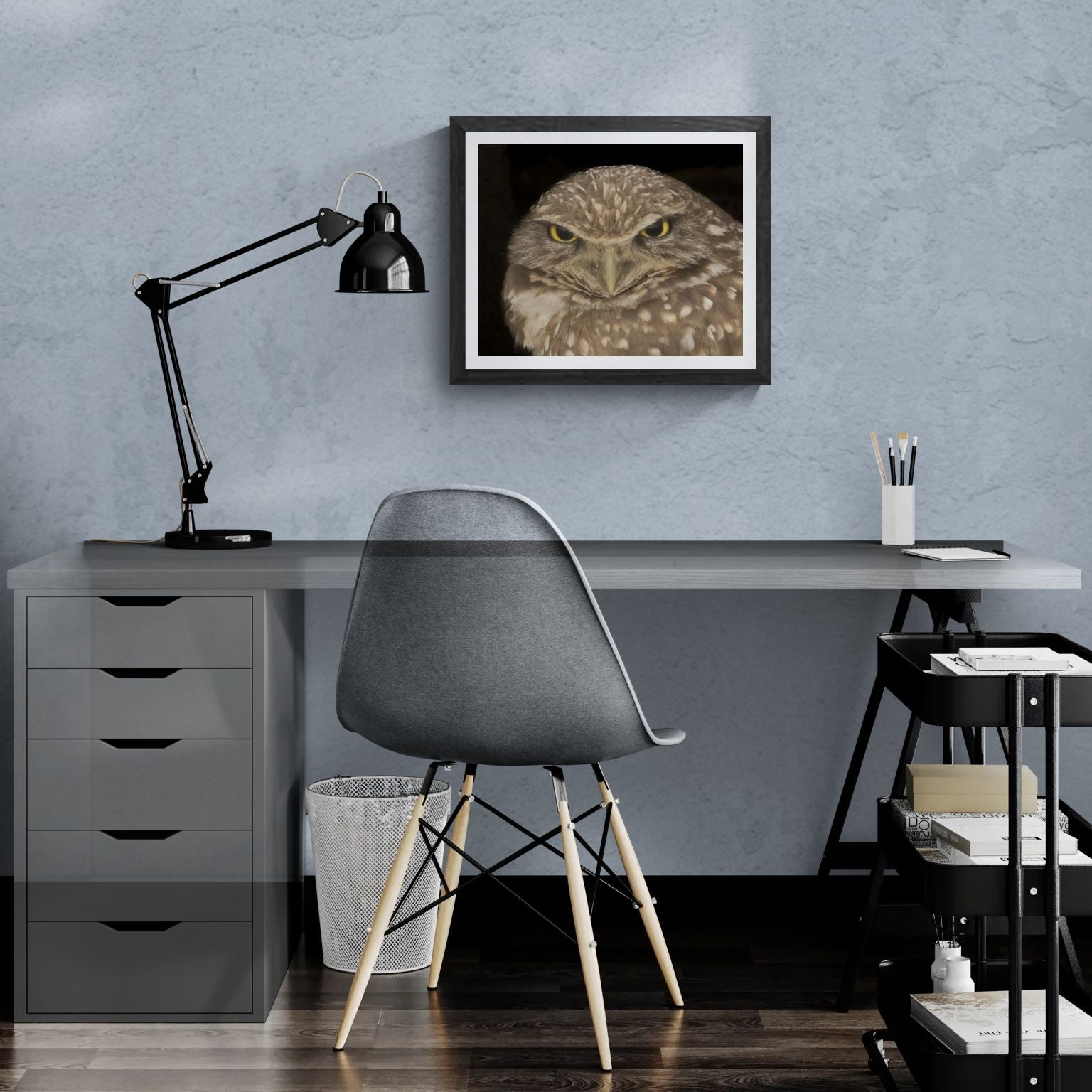 A 20x16 "Standard Edition" of the "Piercing Intensity" Burrowing Owl print featured in a modern home office. The textured black gallery frame anchors the wildlife portrait on a light-blue textured wall above a white desk, illustrating the print’s biological detail and scale for personal galleries.