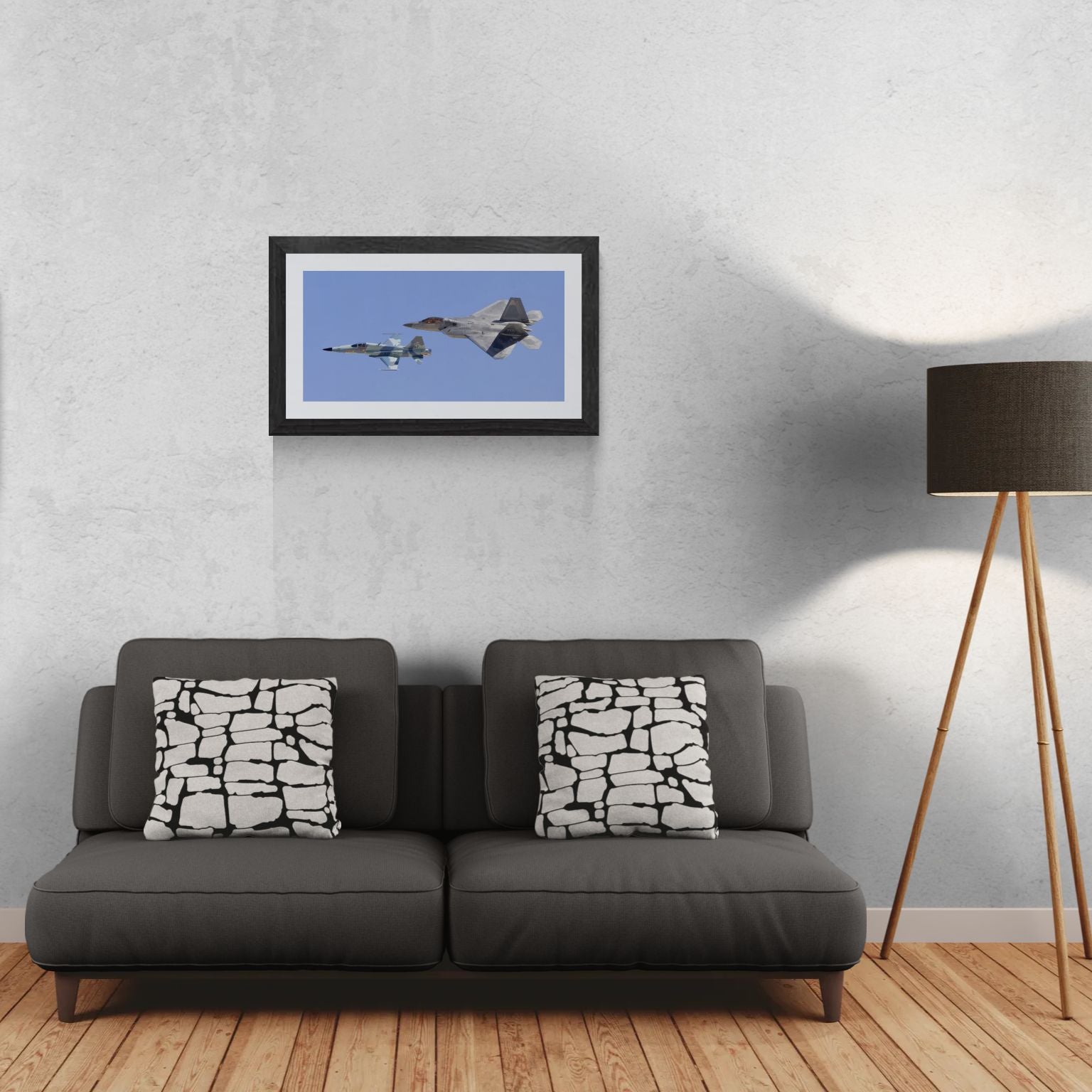 A 30x15 Gallery Edition aviation photograph titled 'Generations,' featuring the historic pairing of an F-22 Raptor and an F-5 Tiger in a Heritage Flight formation. The matted print is displayed in a sleek black frame on a light-textured wall above a modern dark gray sofa. To the right, a tripod floor lamp provides a residential scale reference for this /50 limited edition deployment.