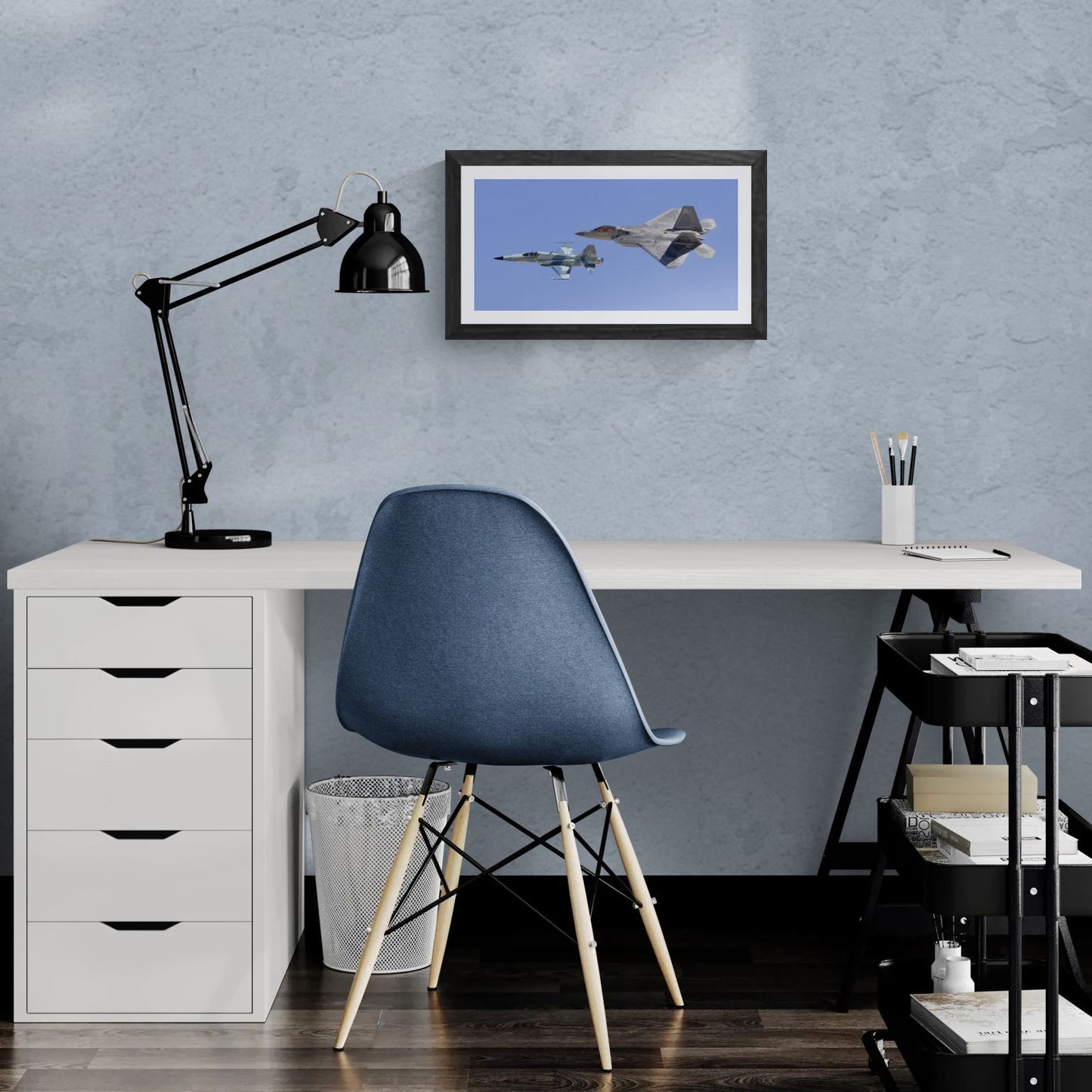 A 20x14 "Standard Edition" of the "Generations" heritage flight print, featuring the F-22 Raptor and F-5 Tiger, displayed in a modern home office. The textured black gallery frame anchors the print on a light-blue textured wall above a white corner desk and a blue ergonomic chair, showcasing its scale for personal collectors.