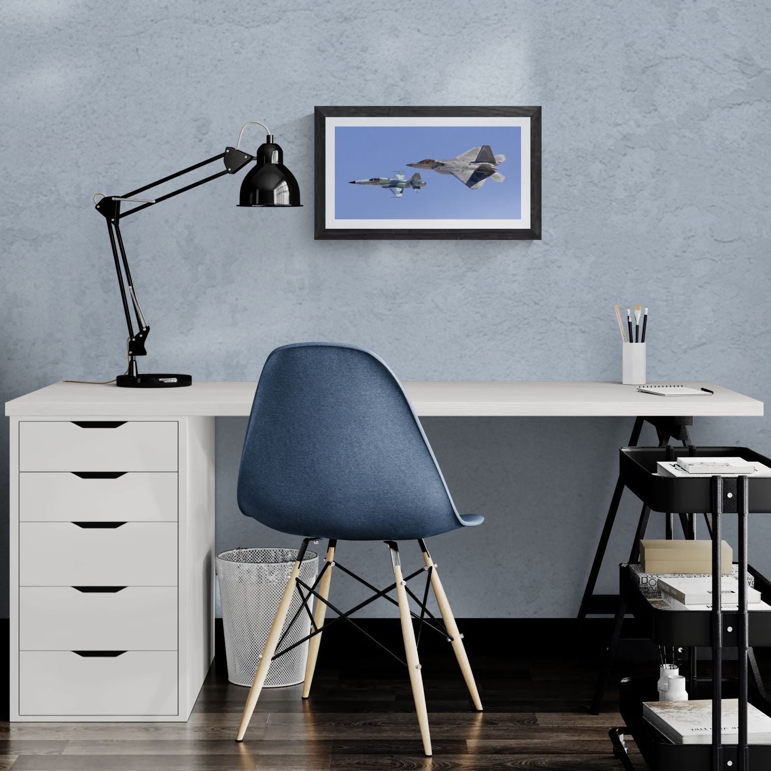 A 20x14 "Standard Edition" of the "Generations" heritage flight print, featuring the F-22 Raptor and F-5 Tiger, displayed in a modern home office. The textured black gallery frame anchors the print on a light-blue textured wall above a white corner desk and a blue ergonomic chair, showcasing its scale for personal collectors.