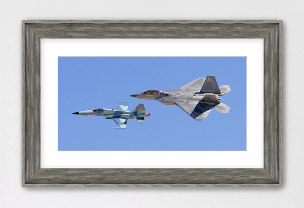 Fine art panoramic print titled "Generations" by PES Editions. The image features a US Air Force F-22 Raptor flying in formation with a camouflaged F-5 Tiger II against a blue sky, showcasing the contrast between stealth technology and classic aviation history.
