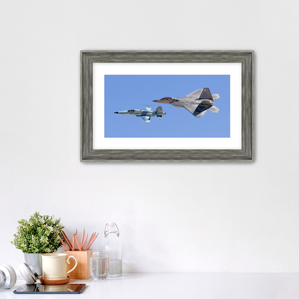 Framed panoramic aviation print titled "Generations" hanging in a modern home office. The limited edition artwork by PES Editions features an F-22 Raptor and F-5 Tiger II flying in formation, presented in a gallery frame to showcase the scale of the panoramic format.