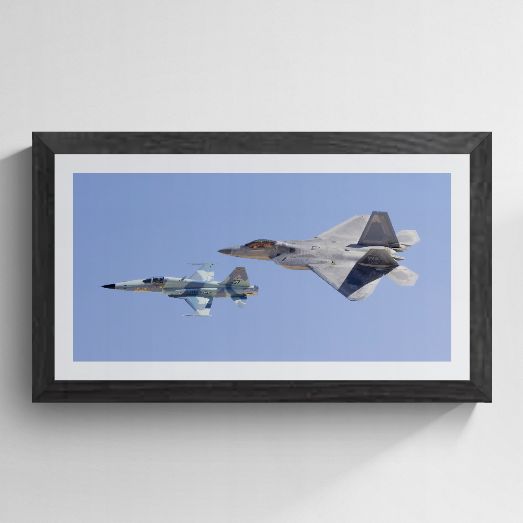 Limited edition fine art photography print titled "Generations" by Phil Schmauder at Nellis AFB. The high-resolution master print, featuring an F-22 Raptor and an F-5 Tiger II in tight heritage formation against a clear blue sky, is professionally presented in a textured black gallery frame with a wide white mat on a clean light grey background.