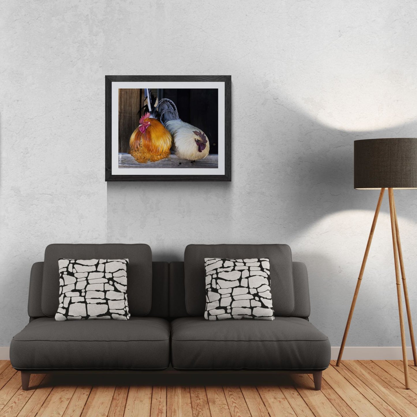 A 30x24 'Gallery Edition' of the 'The Sentinels', featuring a high-resolution barnyard portrait of two roosters with vibrant plumage perched on a weathered wooden beam. The matted print, produced on premium metallic paper to highlight iridescent feather details, is displayed in a textured black gallery frame on a light-textured wall above a modern dark gray sofa. To the right, a tripod floor lamp provides a residential scale reference for this /10 limited edition deployment.