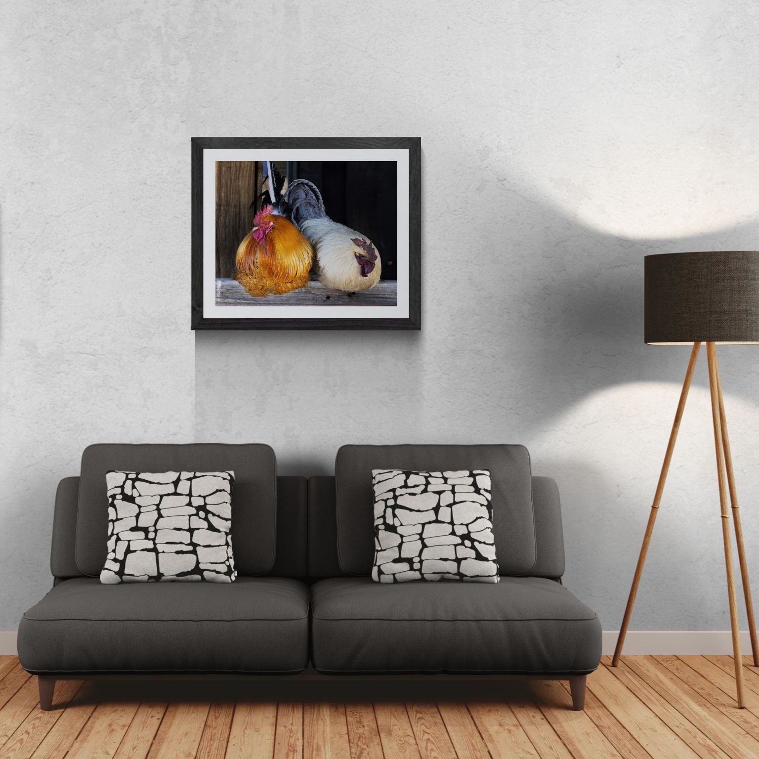 A 30x24 'Gallery Edition' of the 'The Sentinels', featuring a high-resolution barnyard portrait of two roosters with vibrant plumage perched on a weathered wooden beam. The matted print, produced on premium metallic paper to highlight iridescent feather details, is displayed in a textured black gallery frame on a light-textured wall above a modern dark gray sofa. To the right, a tripod floor lamp provides a residential scale reference for this /10 limited edition deployment.