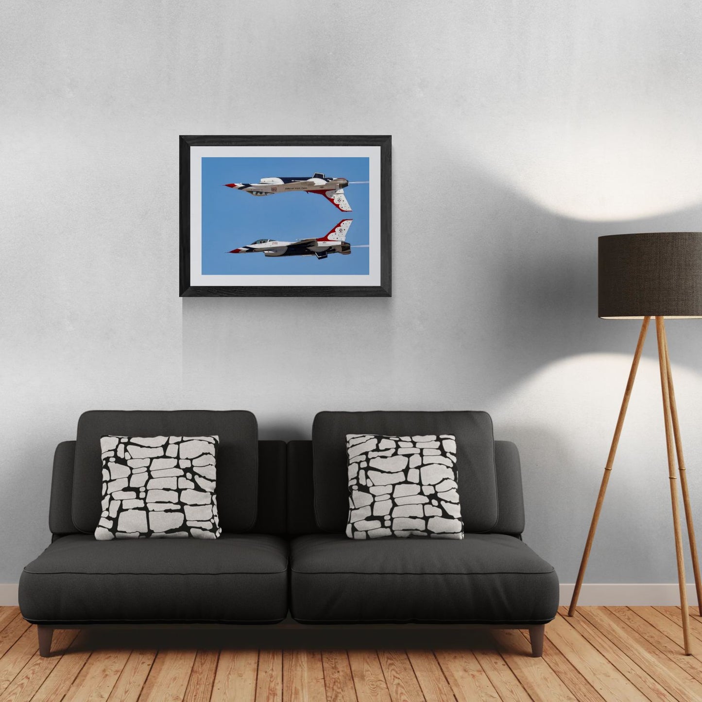 A framed 30x21 Gallery Edition photograph of two U.S. Air Force Thunderbird F-16 Fighting Falcons performing a Calypso Pass, with one aircraft flying inverted directly above the other in a clear blue sky. The matted print is displayed in a sleek black frame on a light-textured wall above a modern dark gray sofa. A tripod floor lamp stands to the right, providing a residential scale reference for the mid-size aviation art deployment.