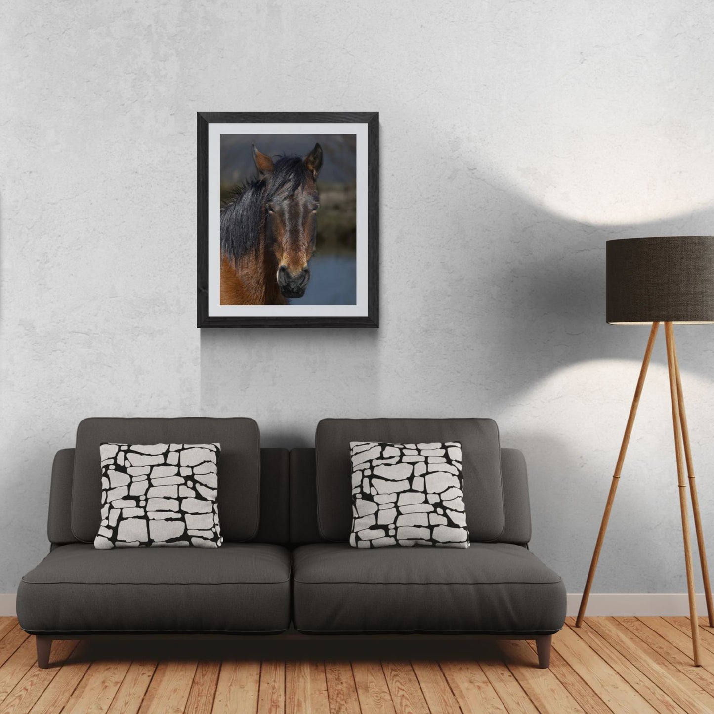A 24x30 vertical 'Gallery Edition' of the 'Wild Mustang: Resilient Spirit' wildlife print, featuring a close-up study of a mustang with a dark, rich coat. The textured black gallery frame anchors the matted print on a light-textured wall above a modern dark gray sofa. To the right, a tripod floor lamp provides a residential scale reference for this /50 limited edition deployment, showcasing the fine archival textures and natural presence of the subject.