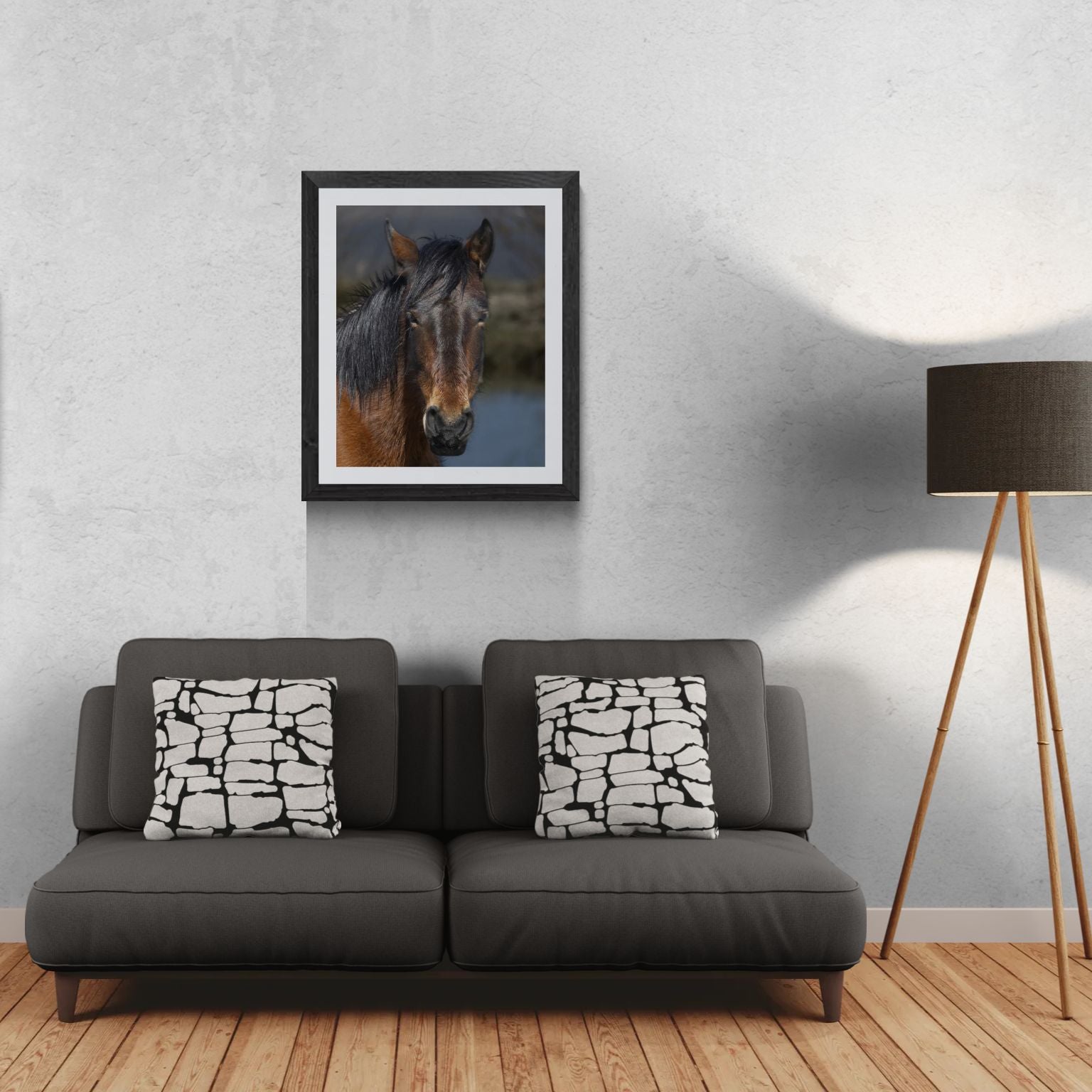 A 24x30 vertical 'Gallery Edition' of the 'Wild Mustang: Resilient Spirit' wildlife print, featuring a close-up study of a mustang with a dark, rich coat. The textured black gallery frame anchors the matted print on a light-textured wall above a modern dark gray sofa. To the right, a tripod floor lamp provides a residential scale reference for this /50 limited edition deployment, showcasing the fine archival textures and natural presence of the subject.