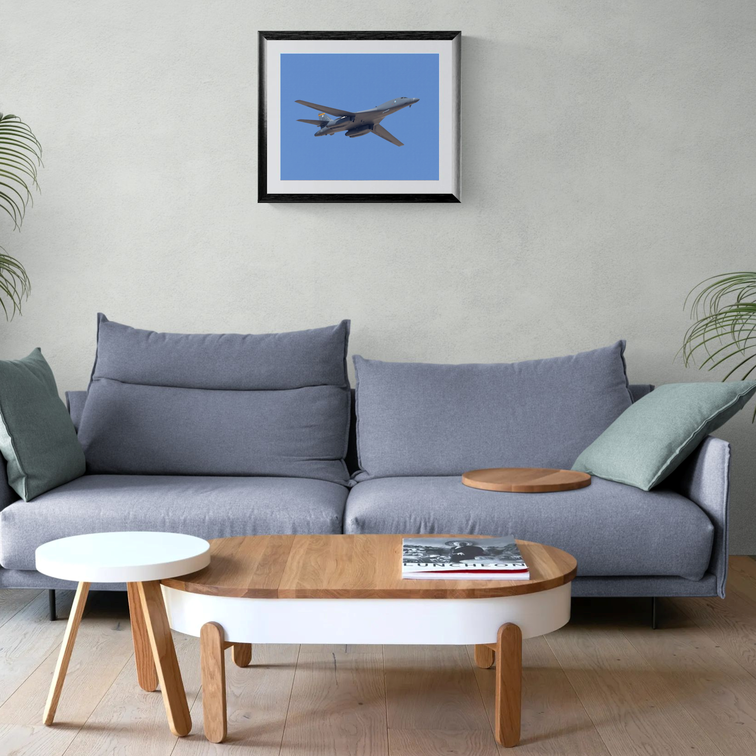 A large-format fine art photography print of a USAF B-1 Lancer bomber jet showcased in a black frame with a clean white mat. The print is centered on a light neutral living room wall directly above a contemporary gray couch with accent pillows.