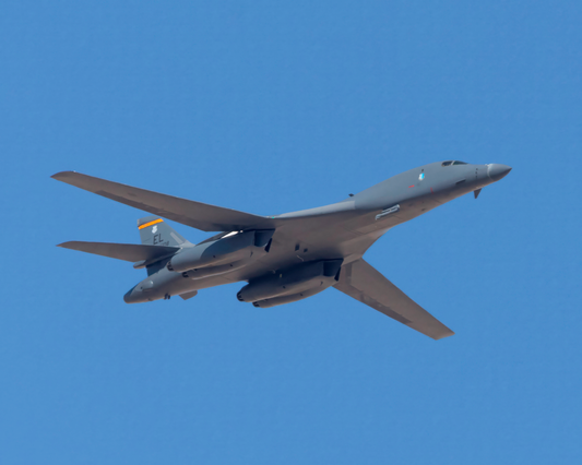 A sharp, full-bleed fine art photograph of a U.S. Air Force B-1B Lancer bomber, affectionately known as The Bone, against a solid, pure blue sky. The underside mechanical details and sweeping wing profile of the supersonic strategic bomber are captured in crisp detail.