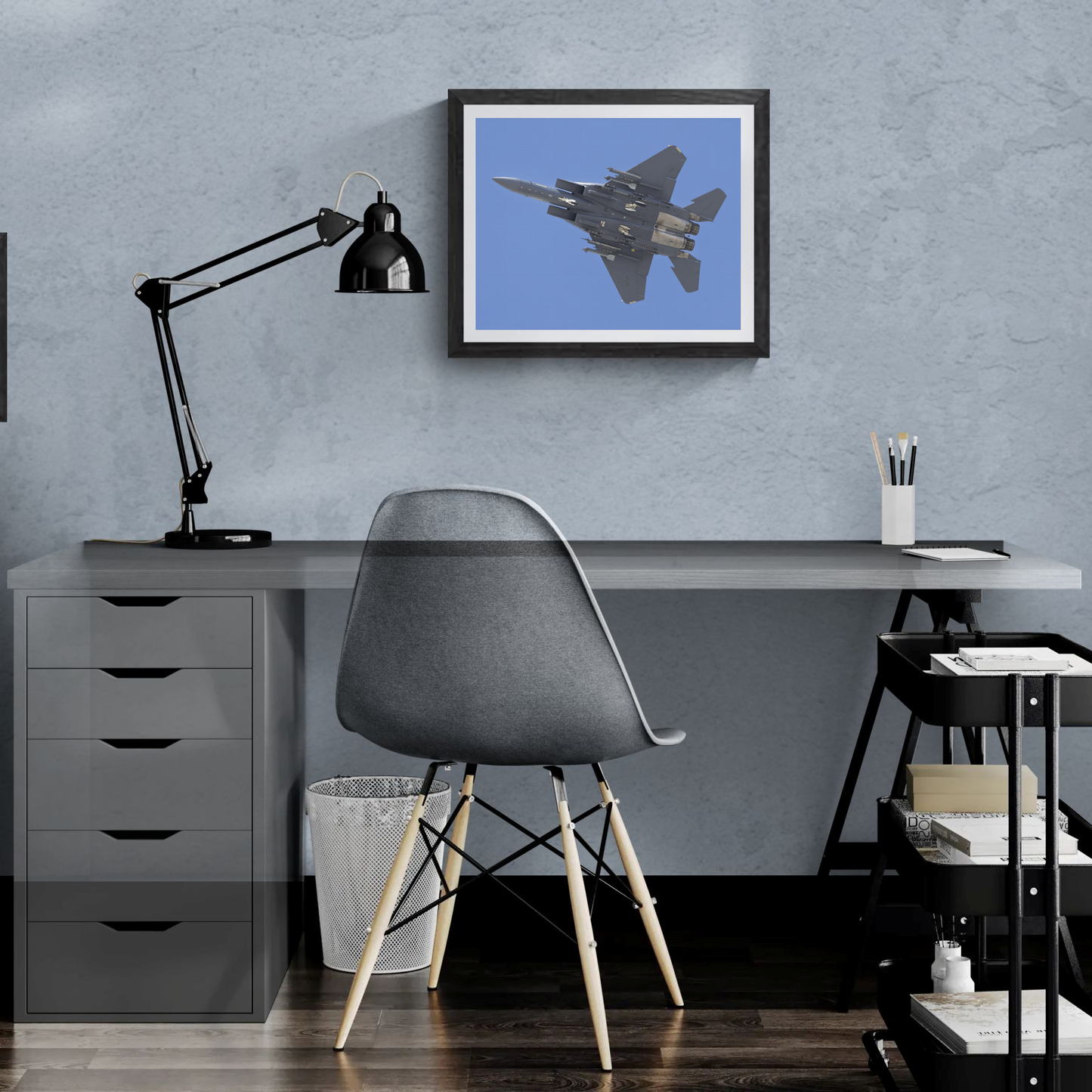 Framed F-15 Strike Eagle print mounted on a slate blue wall above a modern minimalist desk with a task lamp