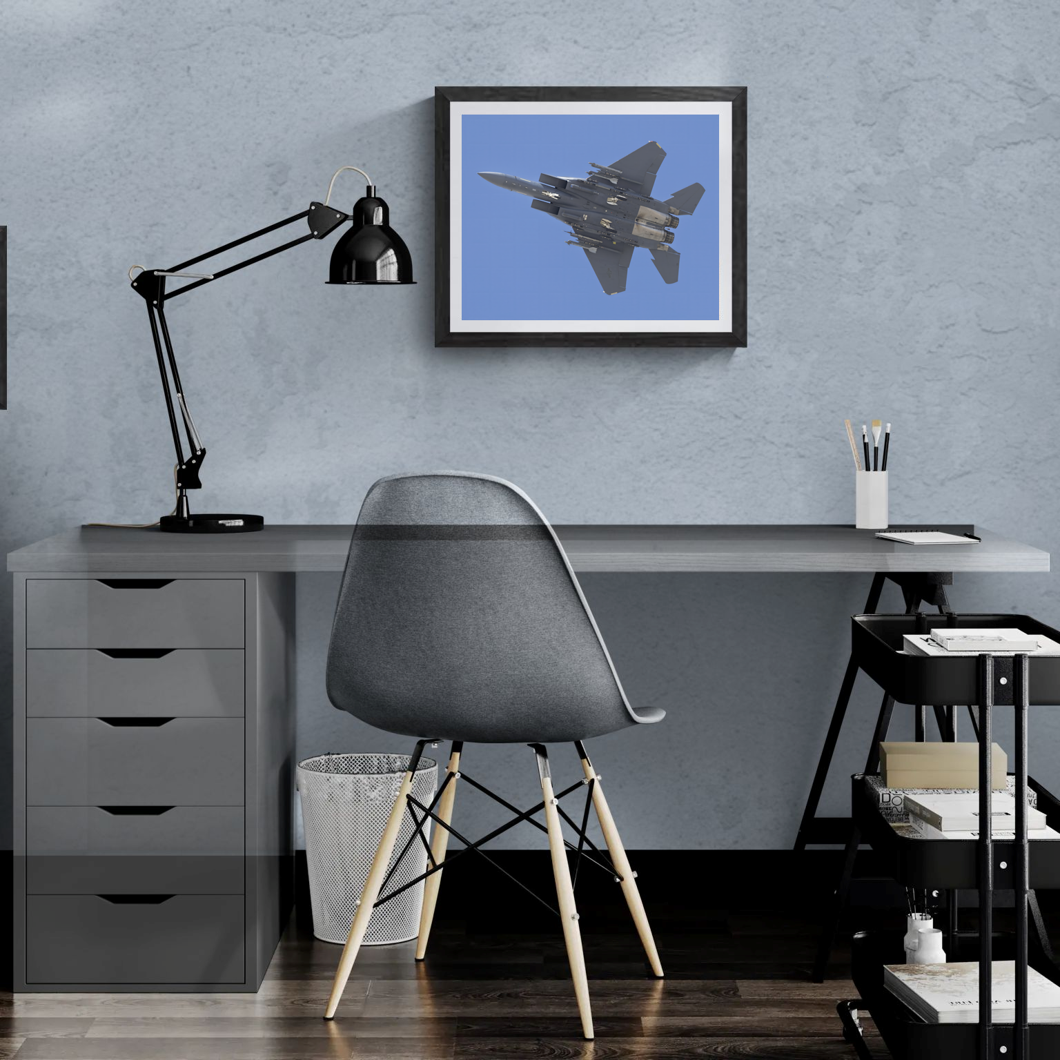 Framed F-15 Strike Eagle print mounted on a slate blue wall above a modern minimalist desk with a task lamp