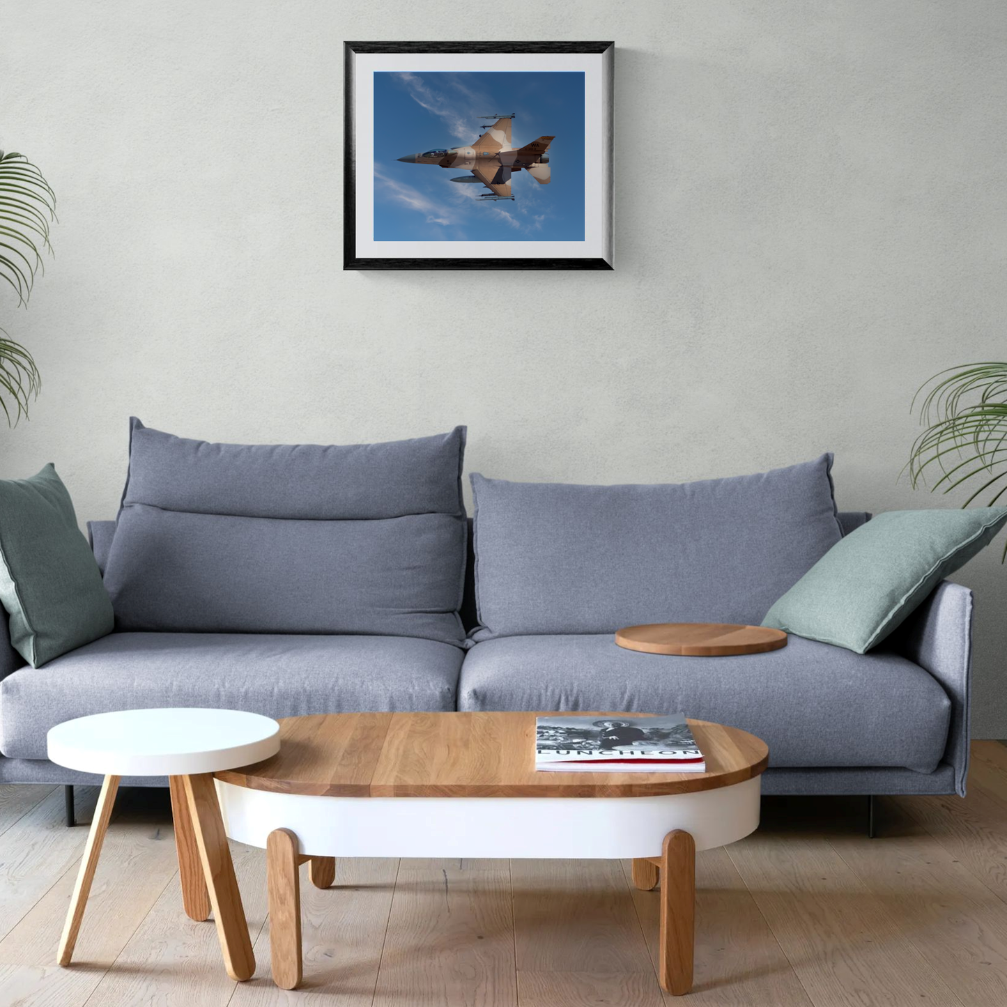A large-format fine art photography print of a desert camo F-16 Aggressor fighter jet, enclosed in a black gallery frame with a clean white mat. The print is centered on a light neutral living room wall directly above a modern gray sofa.