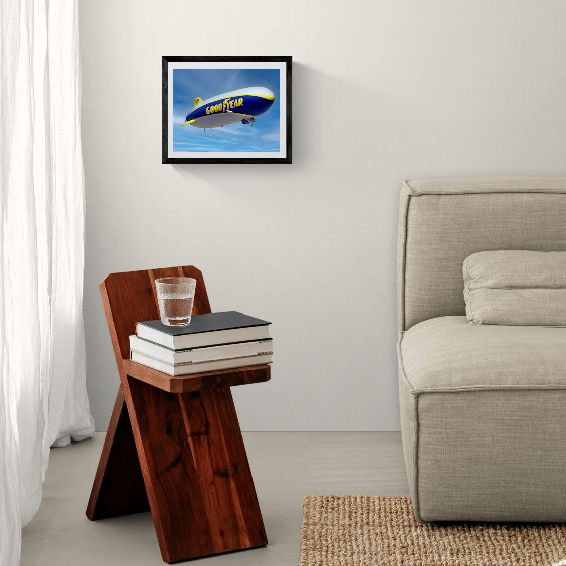 An 11x14 framed fine art print featuring the Goodyear Blimp, Wingfoot Three, against a sweeping blue sky. The minimalist black gallery frame hangs on a crisp white accent wall above a dark wood accent side table next to a neutral-toned couch.