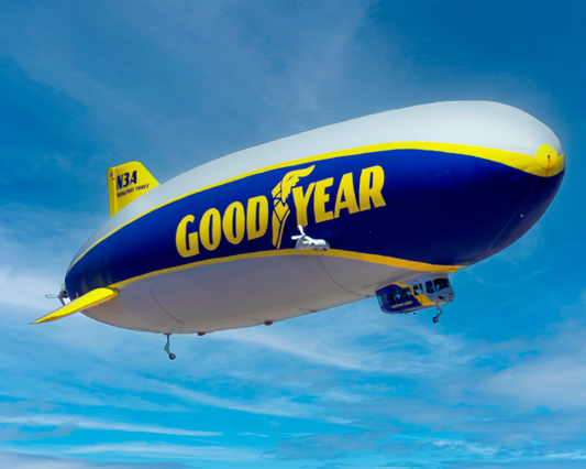 A sharp, full-bleed 10x8 fine art photograph capturing the Goodyear Blimp "Wingfoot Three" soaring smoothly through a clear, vivid blue sky with scattered wispy clouds. The full scale and colorful branding of the iconic semi-rigid airship are visible in crisp detail.