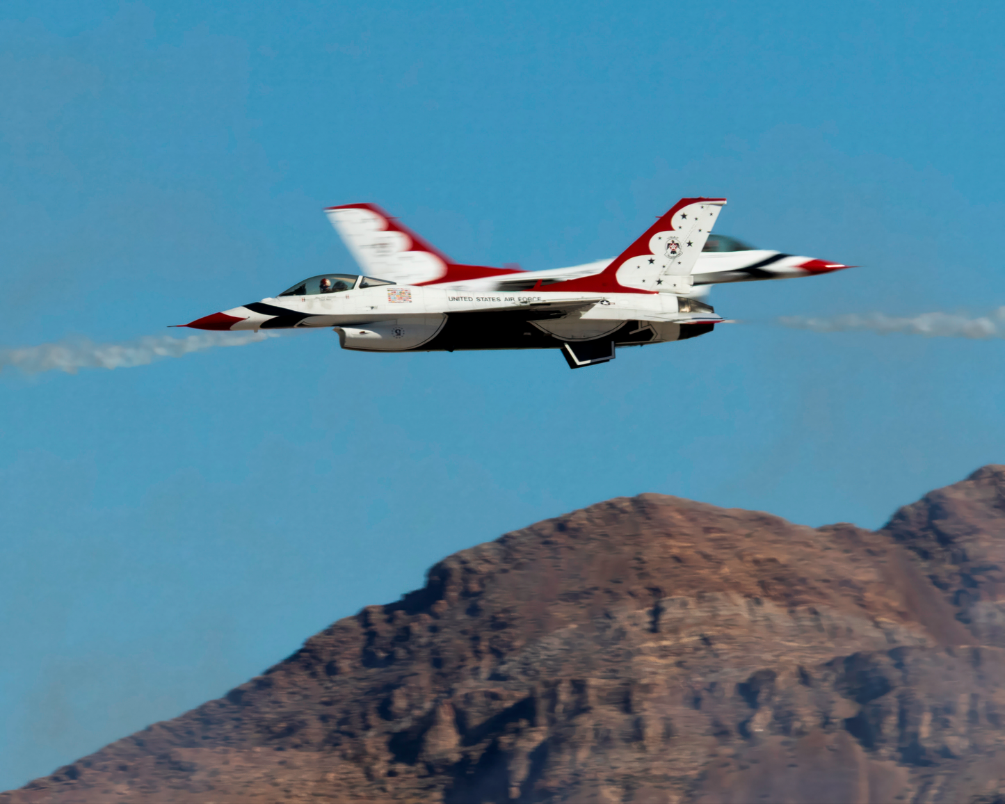 A sharp, high-speed aviation photograph capturing two United States Air Force Thunderbirds F-16 Fighting Falcon jets executing a precise crossover maneuver. The foreground jet is in razor-sharp focus against a clear blue sky, while the rugged, sun-baked ridge line of a desert mountain stands below them, anchoring the incredible aerial display to the American Southwest.