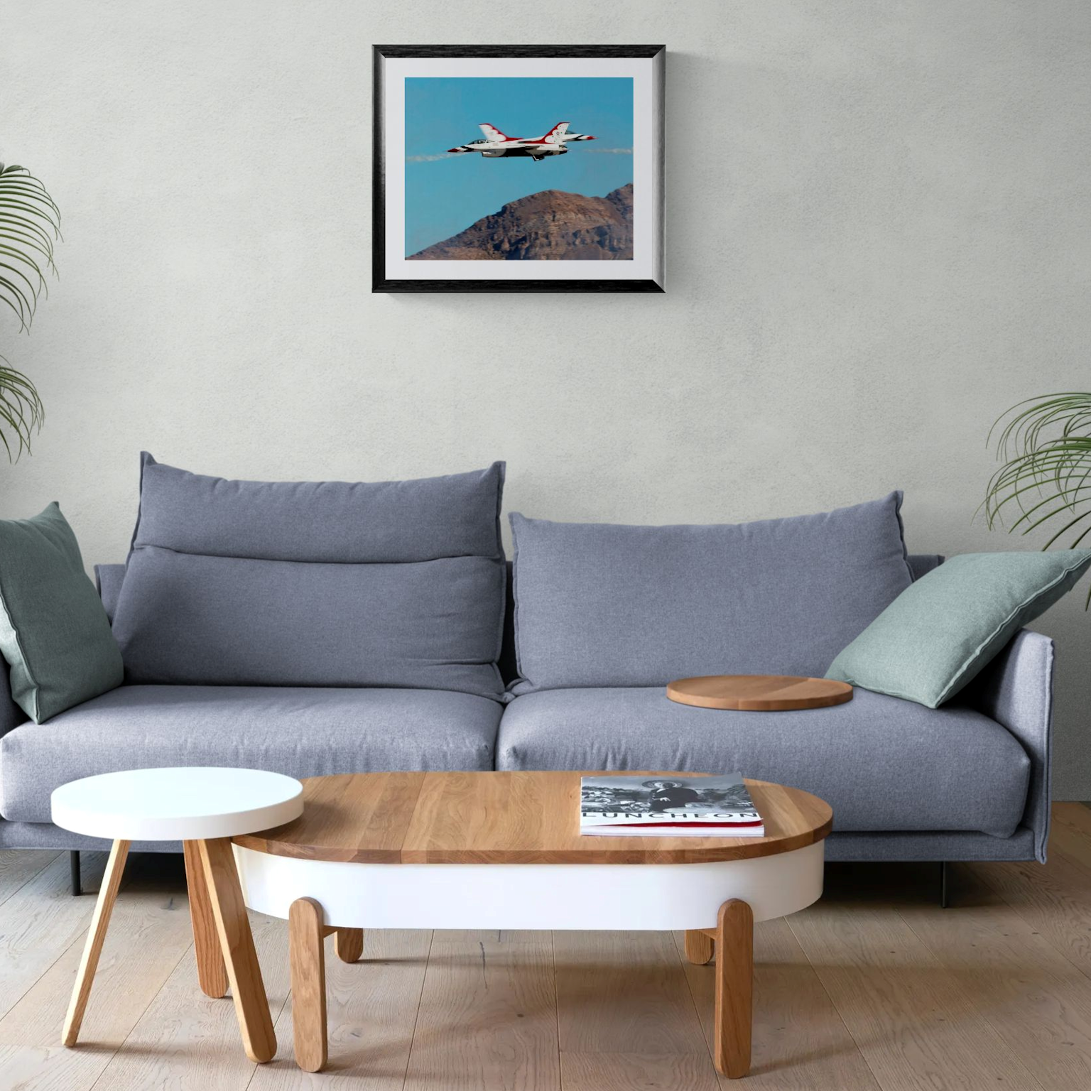 A large-format 16x20 fine art photography print of the USAF Thunderbirds F-16 fighter jets performing a high-speed crossover maneuver against a blue sky and rugged desert mountains. The print is showcased in a black gallery frame with a white mat, centered on a light neutral wall above a contemporary gray sofa with green accent pillows.